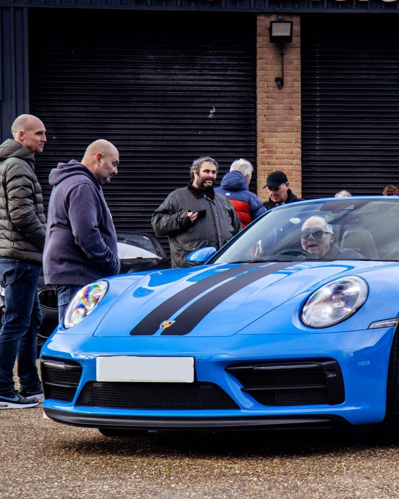 Blue Porsche 911 GTS with people gathered around it in front of a building.